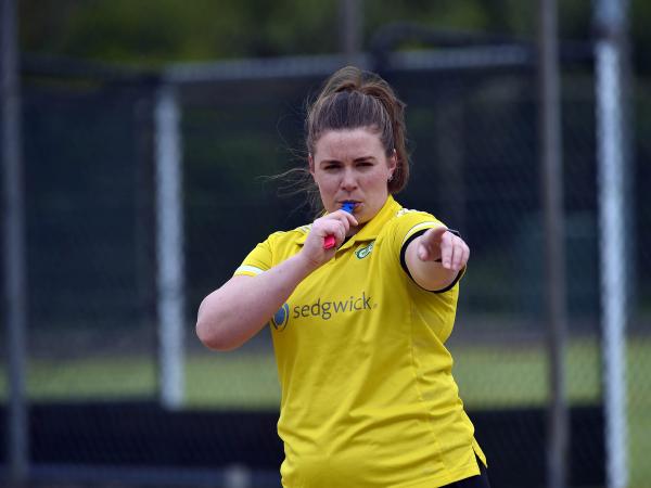 Ellie stand on the pitch, blowing the referee whistle and pointing to the left