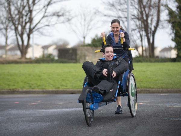 Female runs through the park pushing an adapted wheelchair in front of her with her son