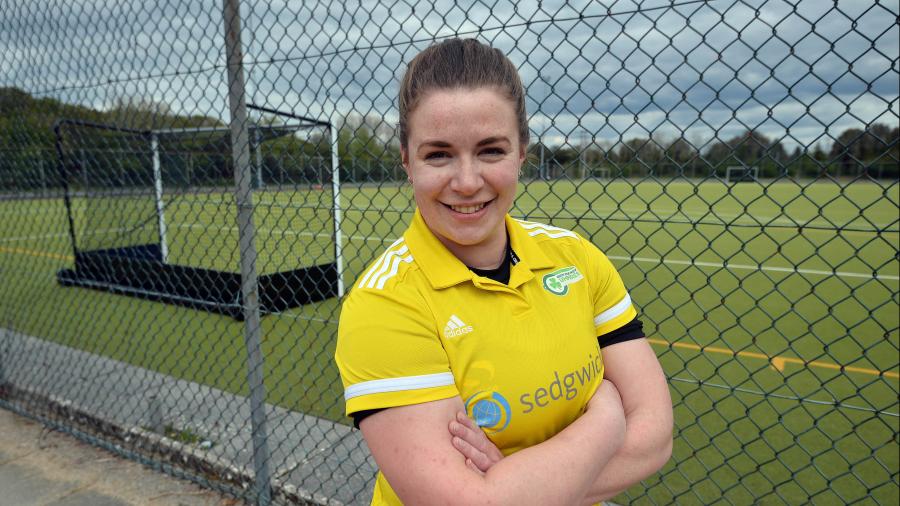 Ellie stands beside a hockey pitch, arms folded wearing a yellow referee shirt