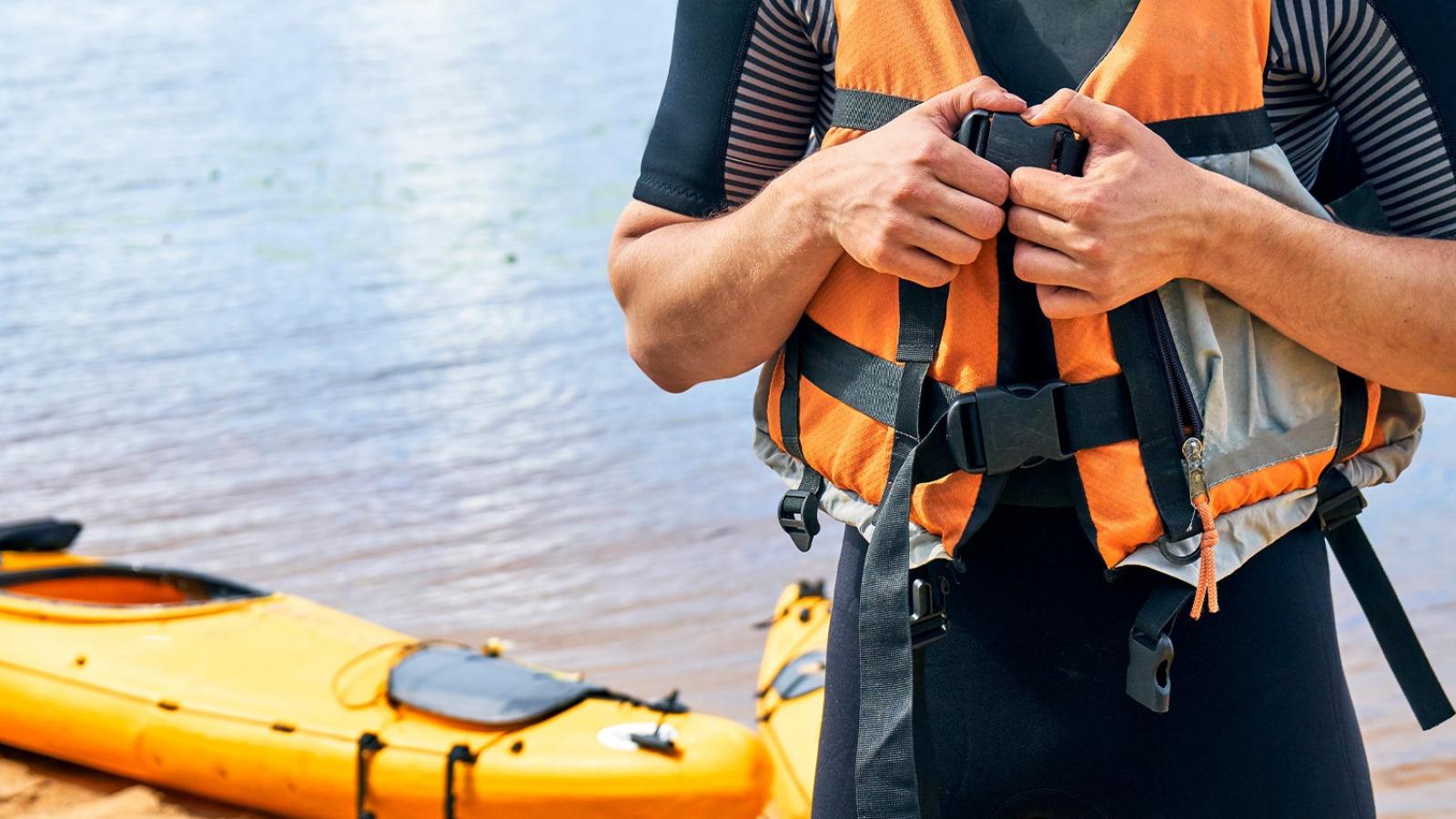 person closes the buckle on a lifejacket with a kayak in the background