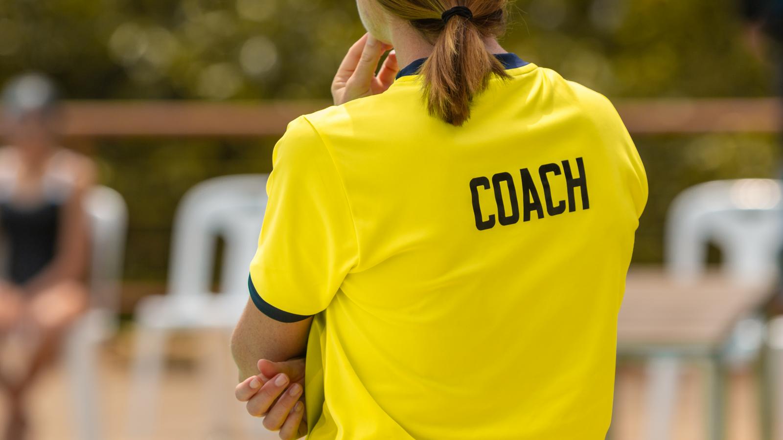 Coach in yellow jersey stands on sideline, background blurred