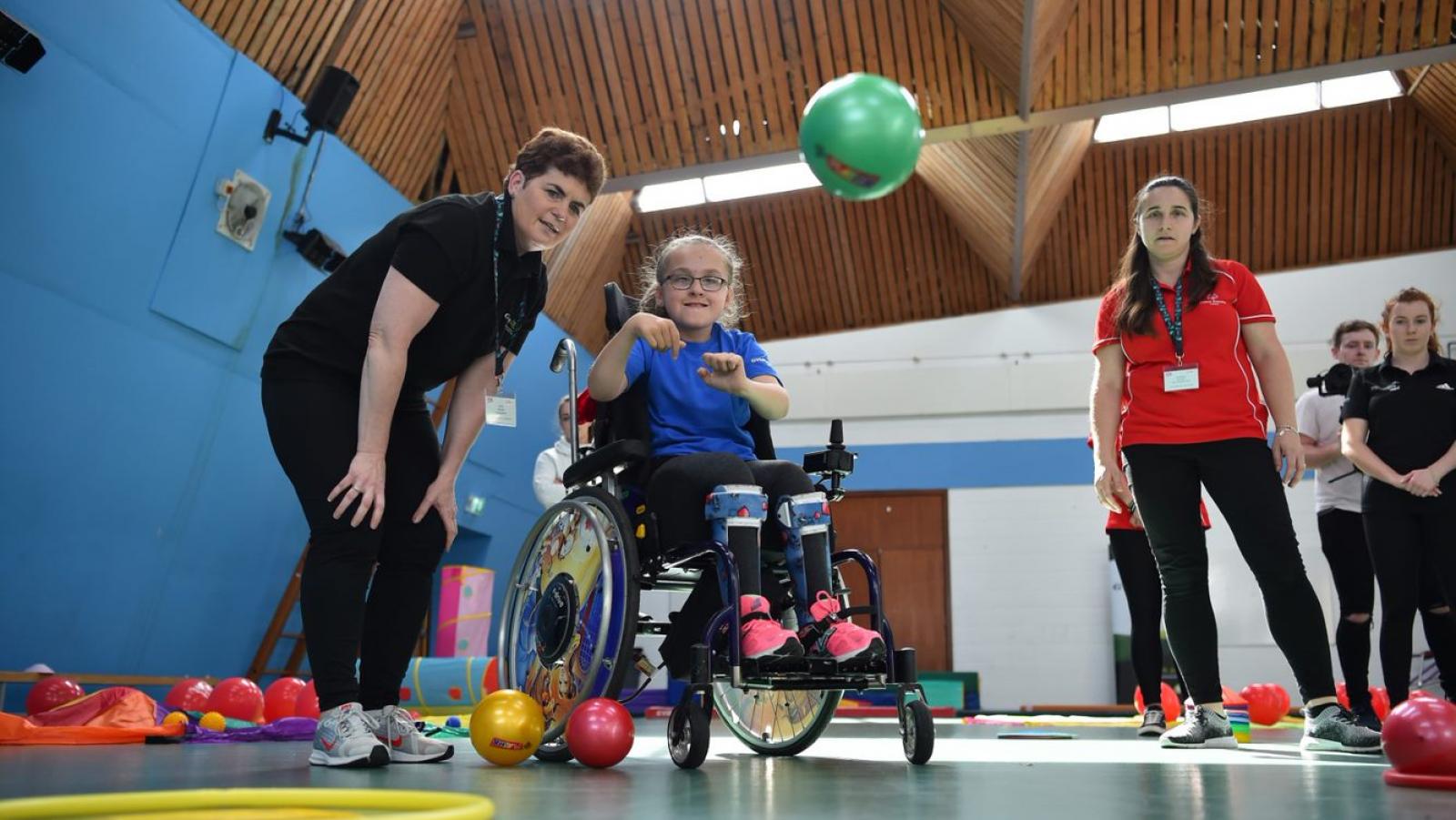 Young girl in a wheelchair throws a green ball as her coach crouches beside her